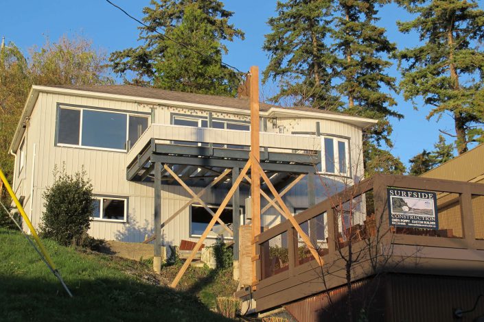 A shot of the old home still standing on the west beach lot ready to be torn down in white rock bc A photo of a crew member guiding in a beam being craned into the lighthouse home in the sooke bc A photo of one of the large, heavy beams being raised into the lighthouse home in sooke bc as built by surfside construction inc.